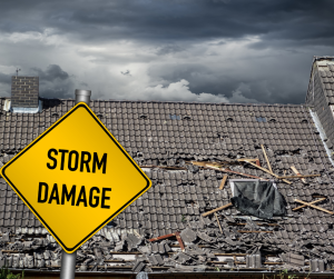 Yellow road sign with black lettering of "Storm Damage". In the background is a gray shingled roof torn up from a recent storm
