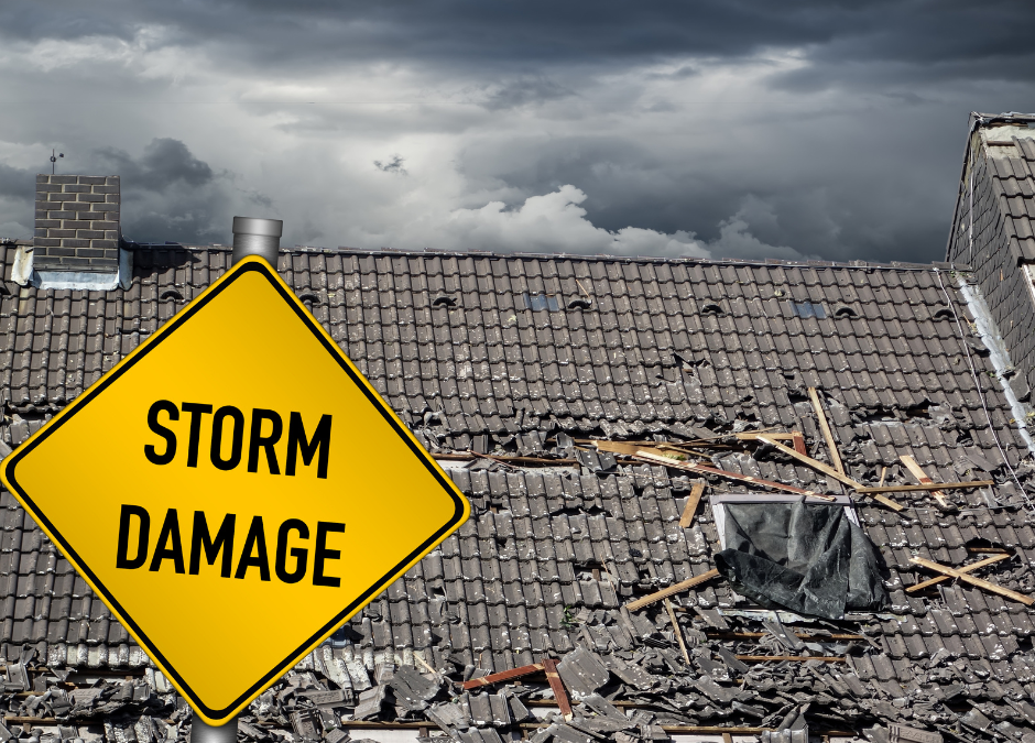 Yellow road sign with black lettering of "Storm Damage". In the background is a gray shingled roof torn up from a recent storm